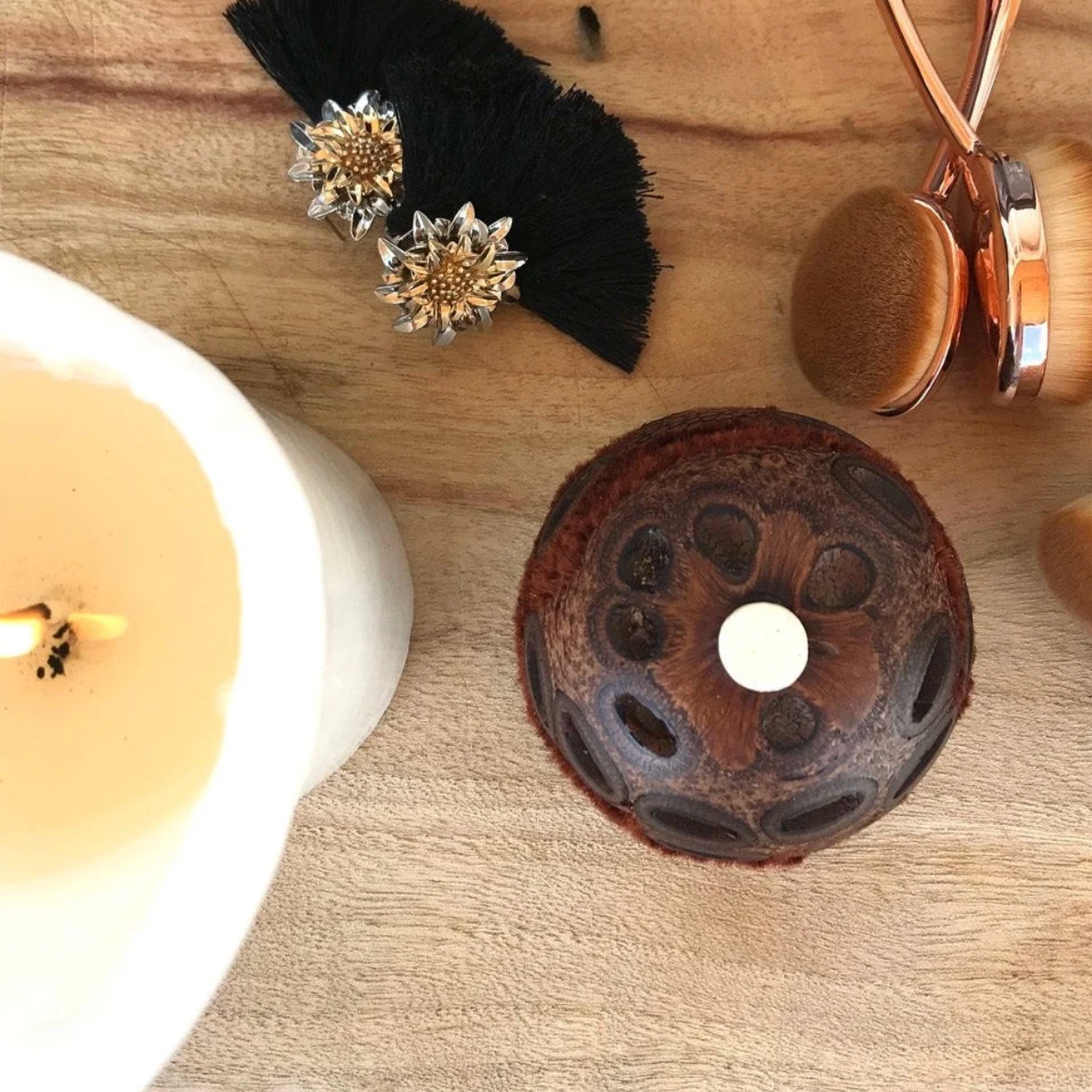 A top-down styled flat-lay on a light wood surface featuring a dark brown lotus seed pod centerpiece with a white wax center, a white candle partially visible on the left, a black feather accessory with two silver floral studs, and a rose gold makeup sponge.