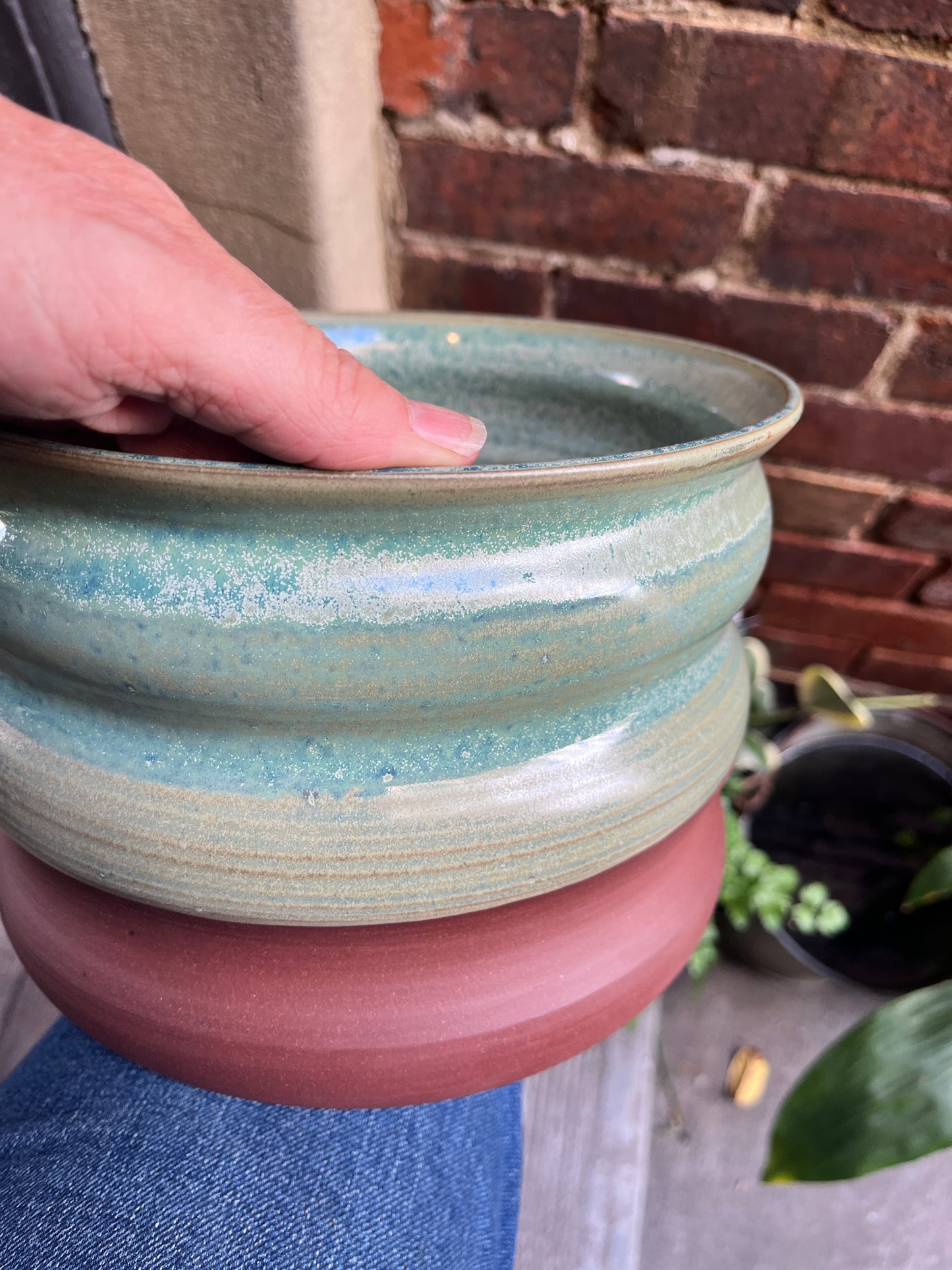Curved ceramic pot of variegated shades of green glaze held by a person against a brick wall.