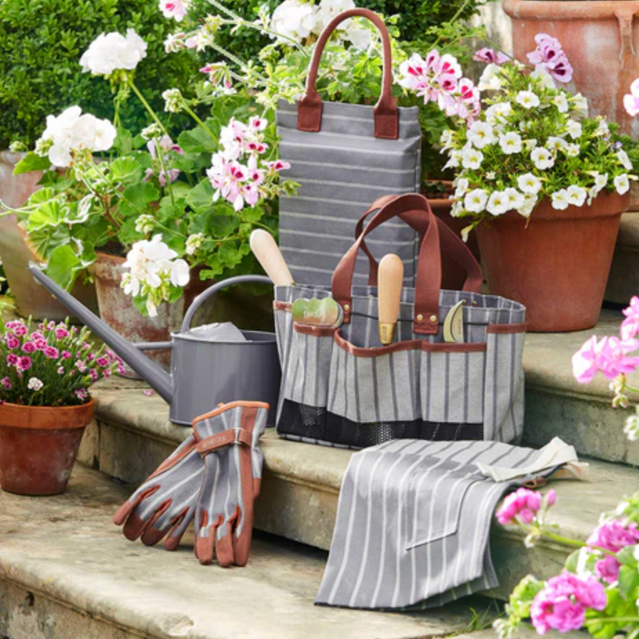 Gardening tools and accessories including a tote bag, gloves, and a watering can on a garden bench with flowers.