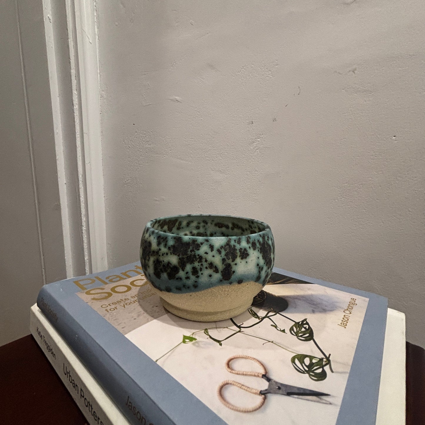 A handmade ceramic bowl with a speckled teal and cream 'moonscape' glaze sits on top of two stacked books against a textured off-white wall.
