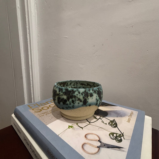 A handmade ceramic bowl with a speckled teal and cream 'moonscape' glaze sits on top of two stacked books against a textured off-white wall.