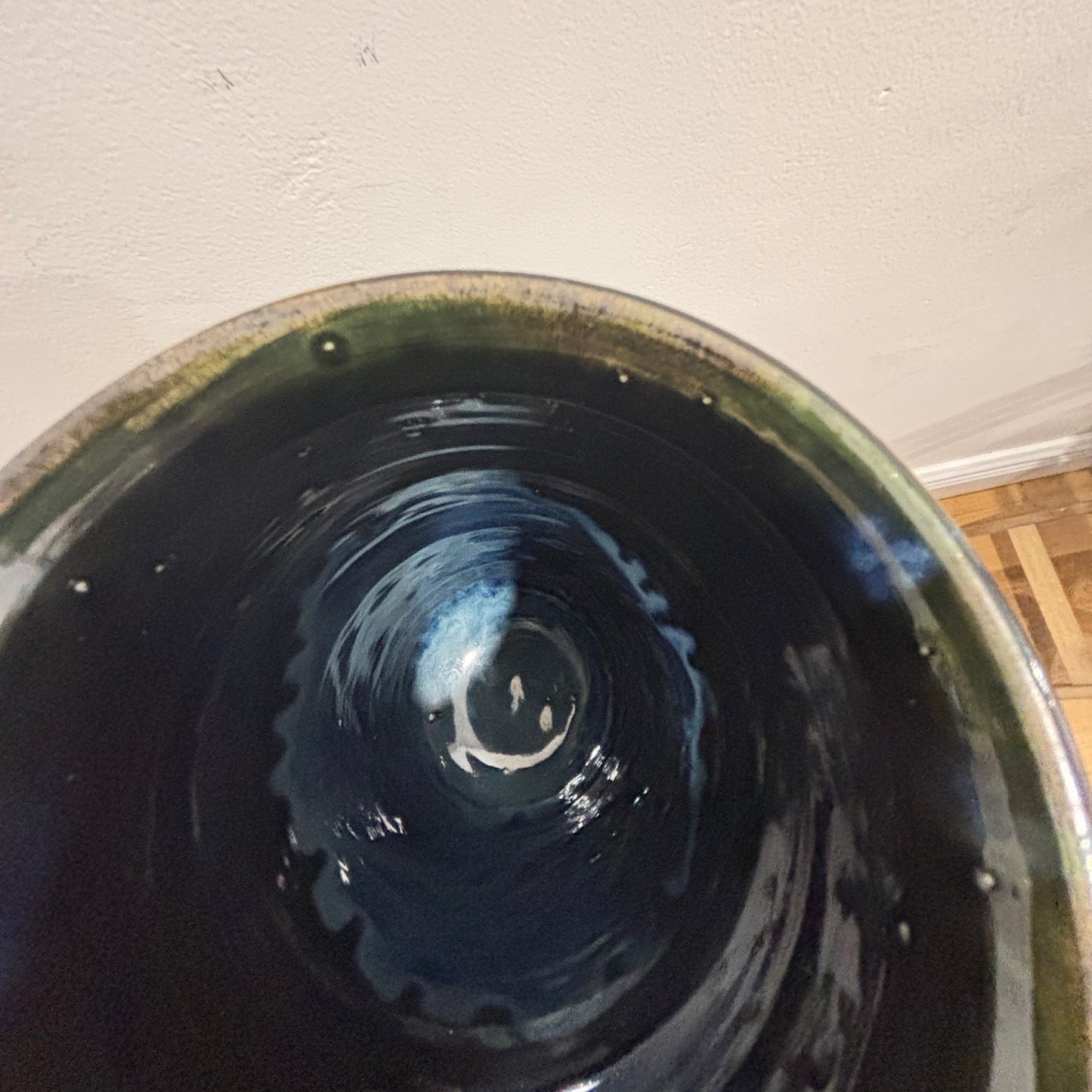 A close-up view from inside a glossy ceramic vase, showing a dark blue and black interior with a green glazed rim, set against a plain off-white wall.