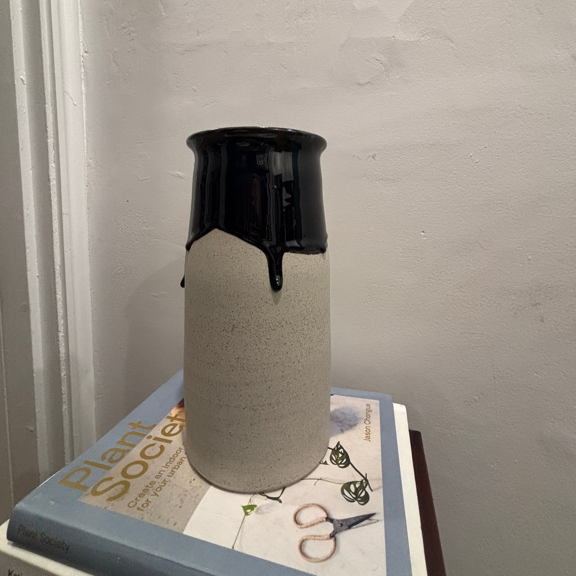 A ceramic vase with a textured light gray body and a glossy blue-black glazed top, displayed on top of books against a plain wall.