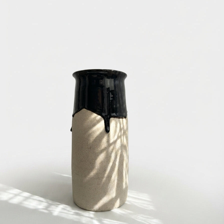 A tall cylindrical ceramic vase with a glossy blue-black glazed top and a matte beige textured lower body, standing on a white surface with soft leaf shadows cast across it.