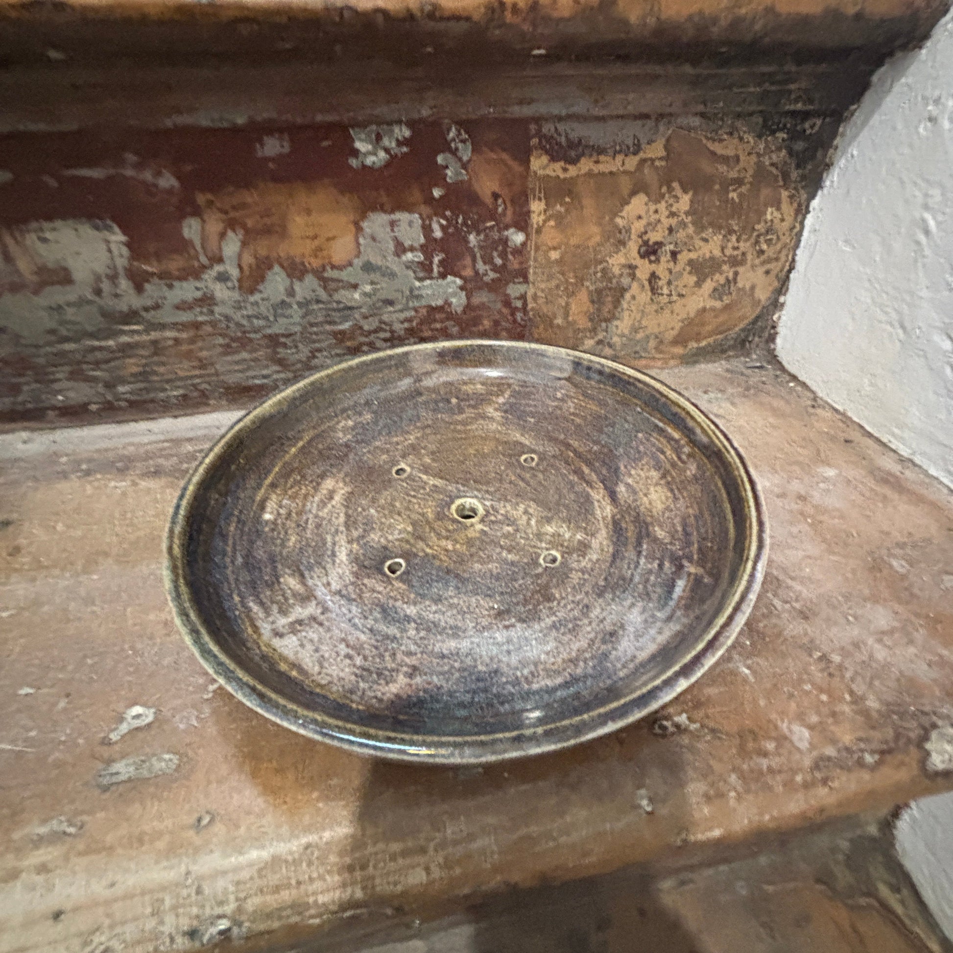 A round, distressed ceramic bonsai planter with a dark brown and blue finish, set on a rustic wooden step against a weathered wall.