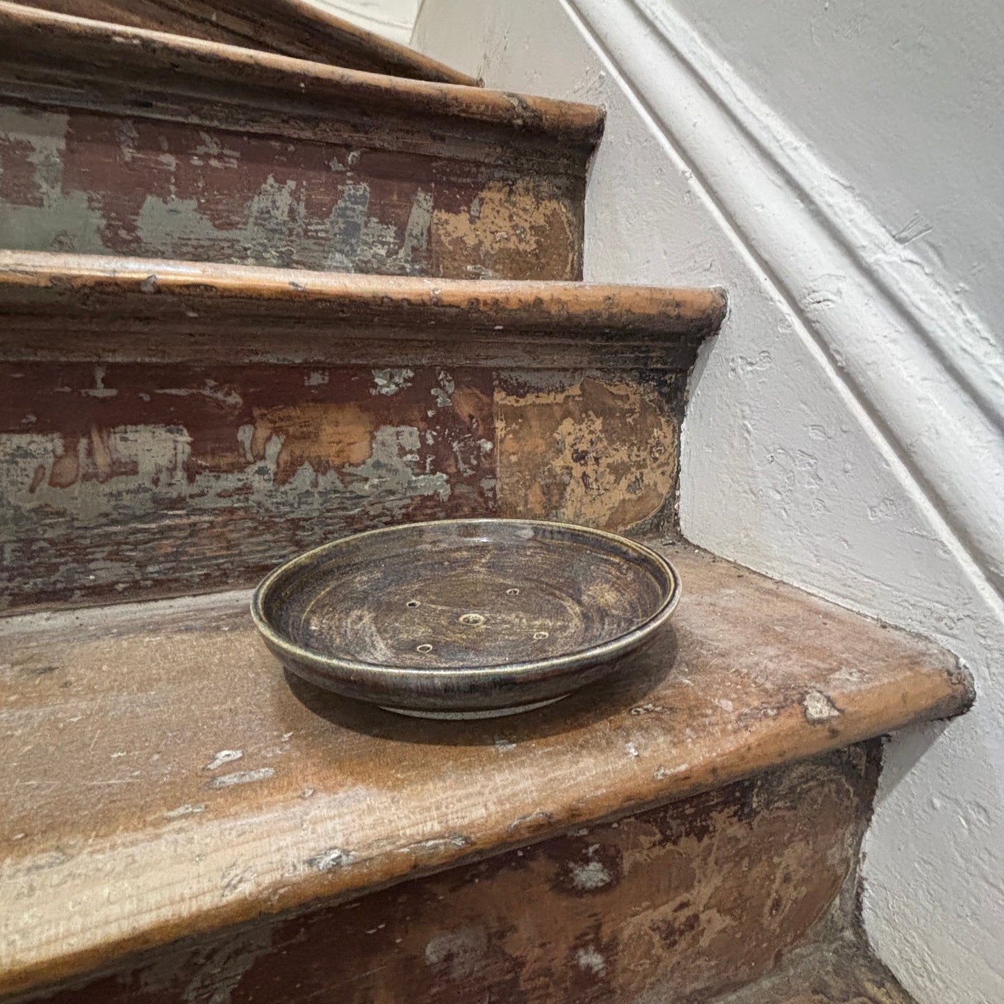 A round, dark ceramic shallow bonsai planter  resting on a weathered wooden staircase with peeling paint and a white wall in the background.