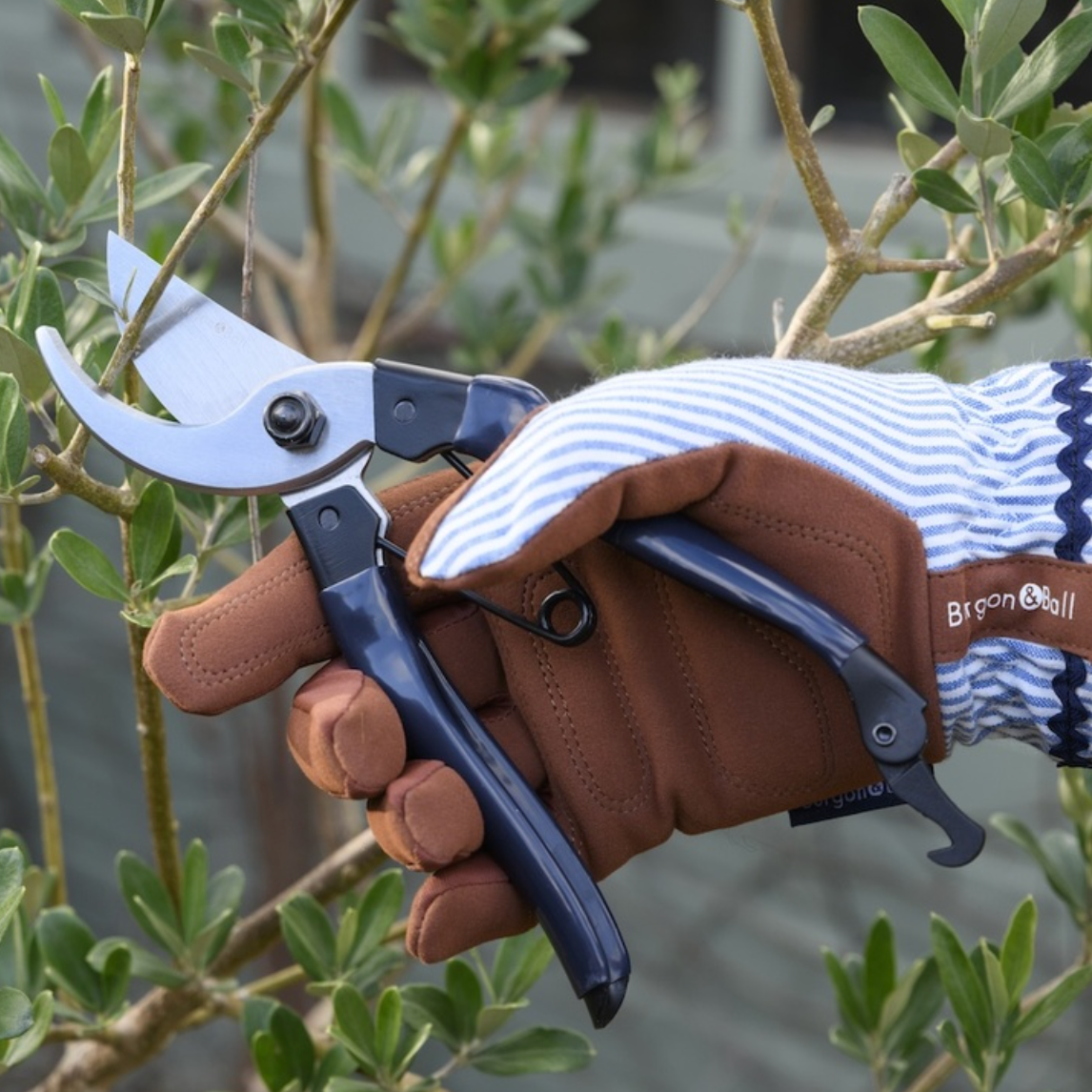 A hand wearing a brown leather gardening glove and a blue-and-white striped knit sleeve holds pruning shears with silver blades and dark blue handles, cutting a thin branch in a garden setting with green leaves and soft gray background.