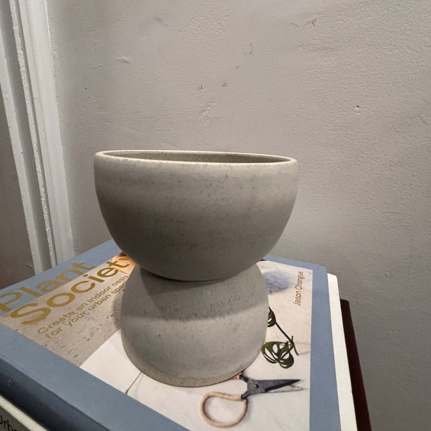 A matte light white/grey ceramic vase with a flared top and bulbous base, displayed on top of books against a textured grey wall.