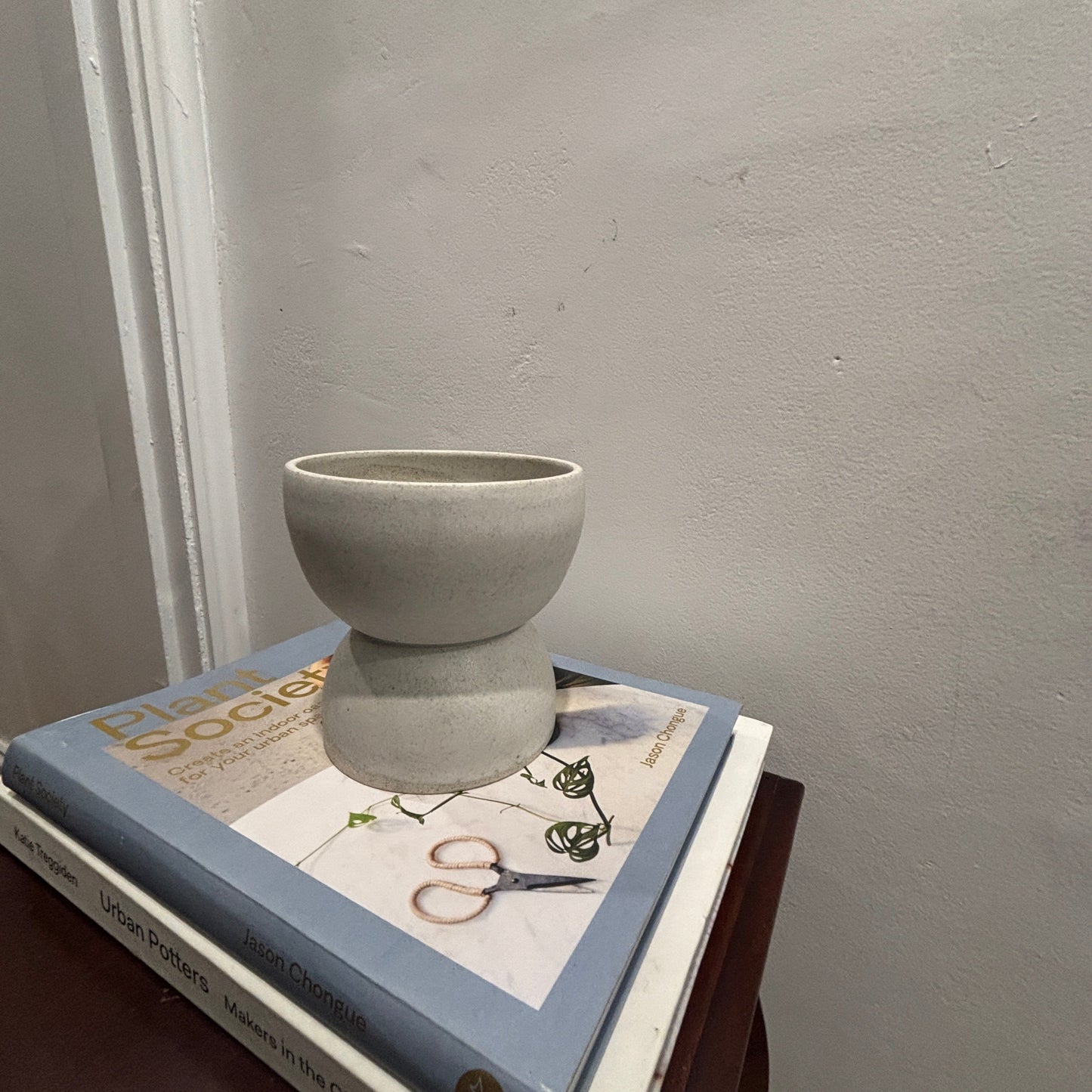 A small matte white/grey ceramic vase with a flared, tiered design sits on top of a stack of books on a dark wooden table against a textured light grey wall.