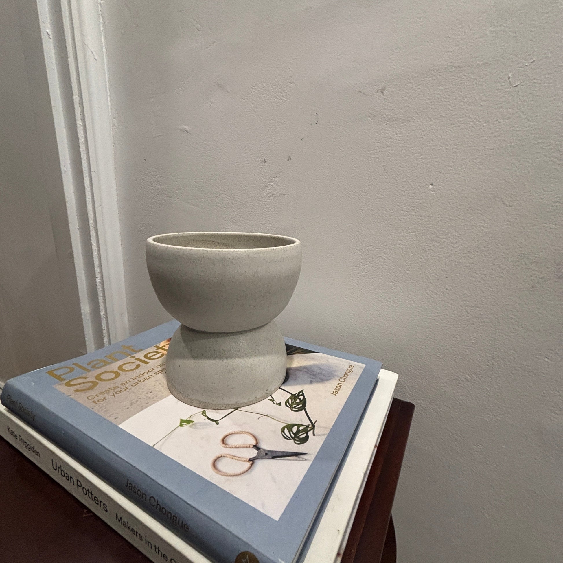A small matte white/grey ceramic vase with a flared, tiered design sits on top of a stack of books on a dark wooden table against a textured light grey wall.