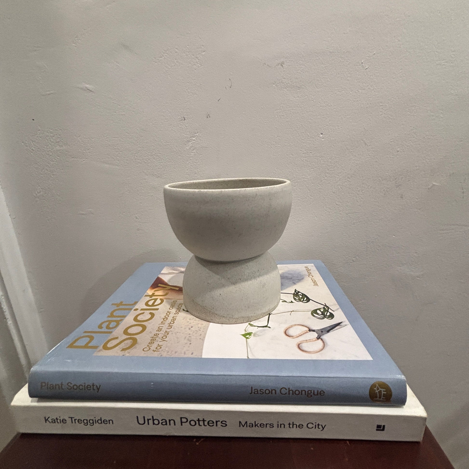 A small matte ceramic vase with a flared pedestal base sits on top of two books against a light grey wall.