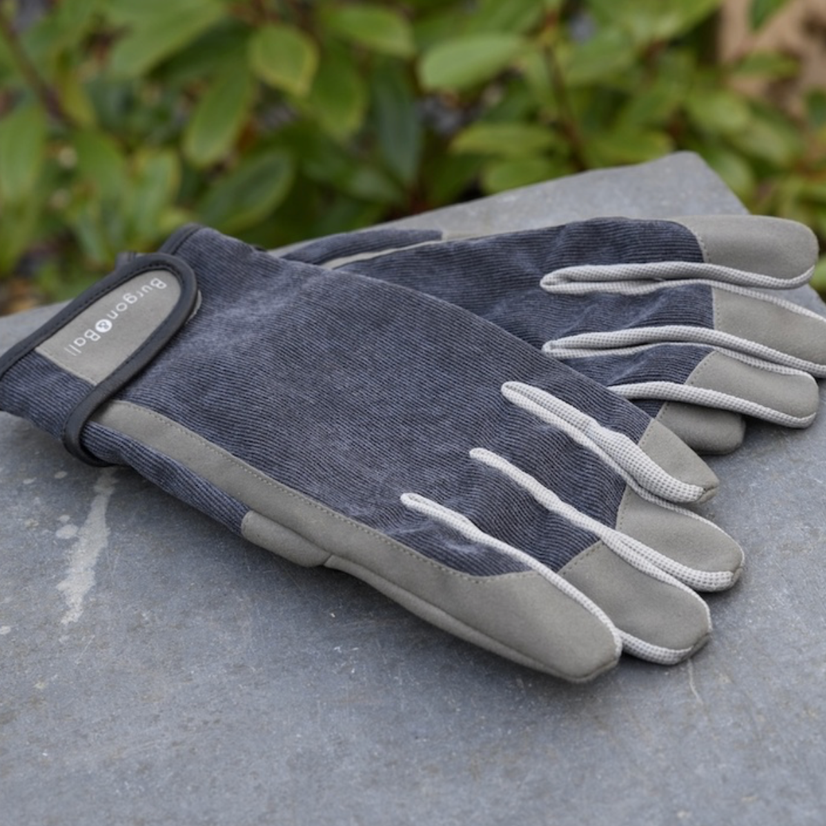 A pair of dark grey work gloves with light grey fingertips and palms, resting on a grey stone surface with blurred green foliage in the background. The brand name 'Burgon + Ball' is visible on the wrist strap.