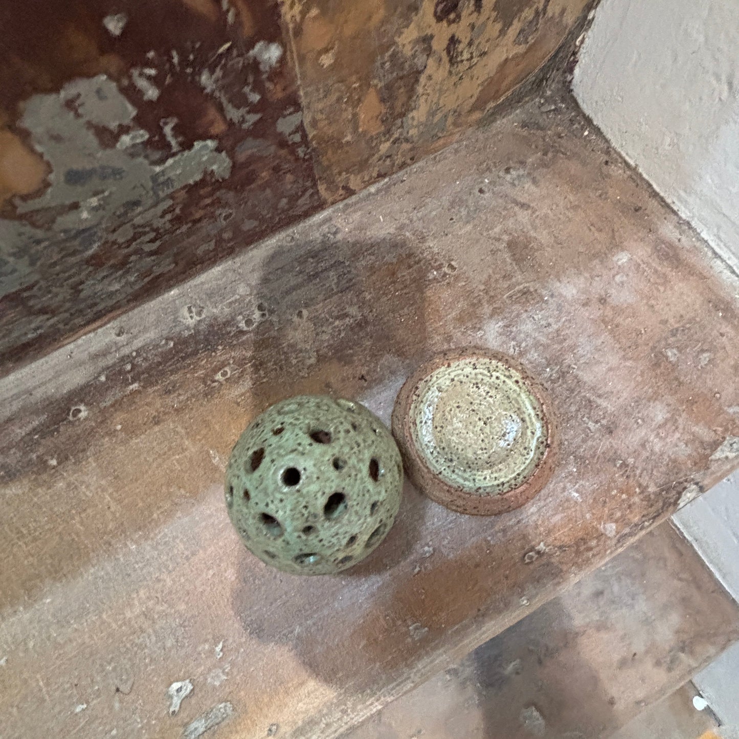 A small ceramic egg shaped incense holder on a rustic wooden shelf: a perforated green-gray sphere and a small round bowl with a speckled beige and brown glaze, set against a weathered wood background.