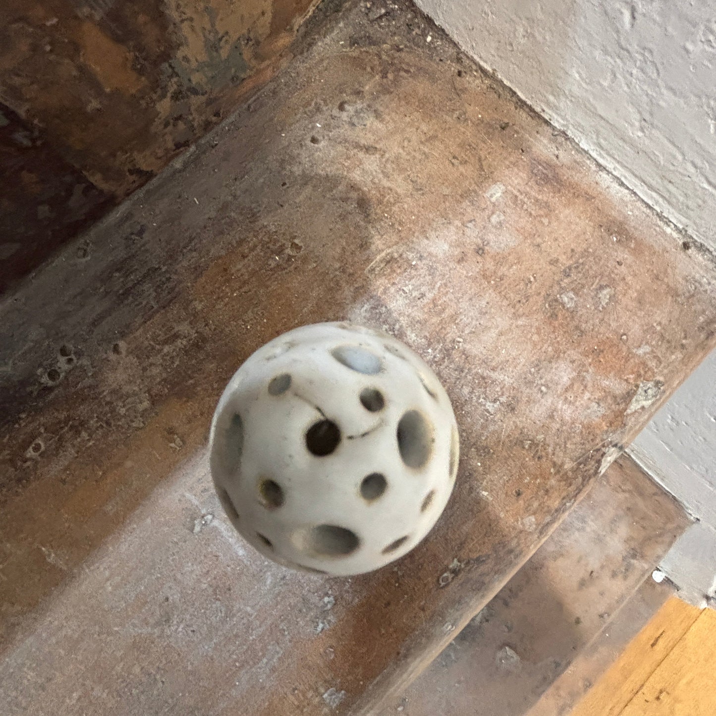 A white ceramic egg shaped incense holder with multiple dark circular holes, resting on a rustic wooden staircase step.
