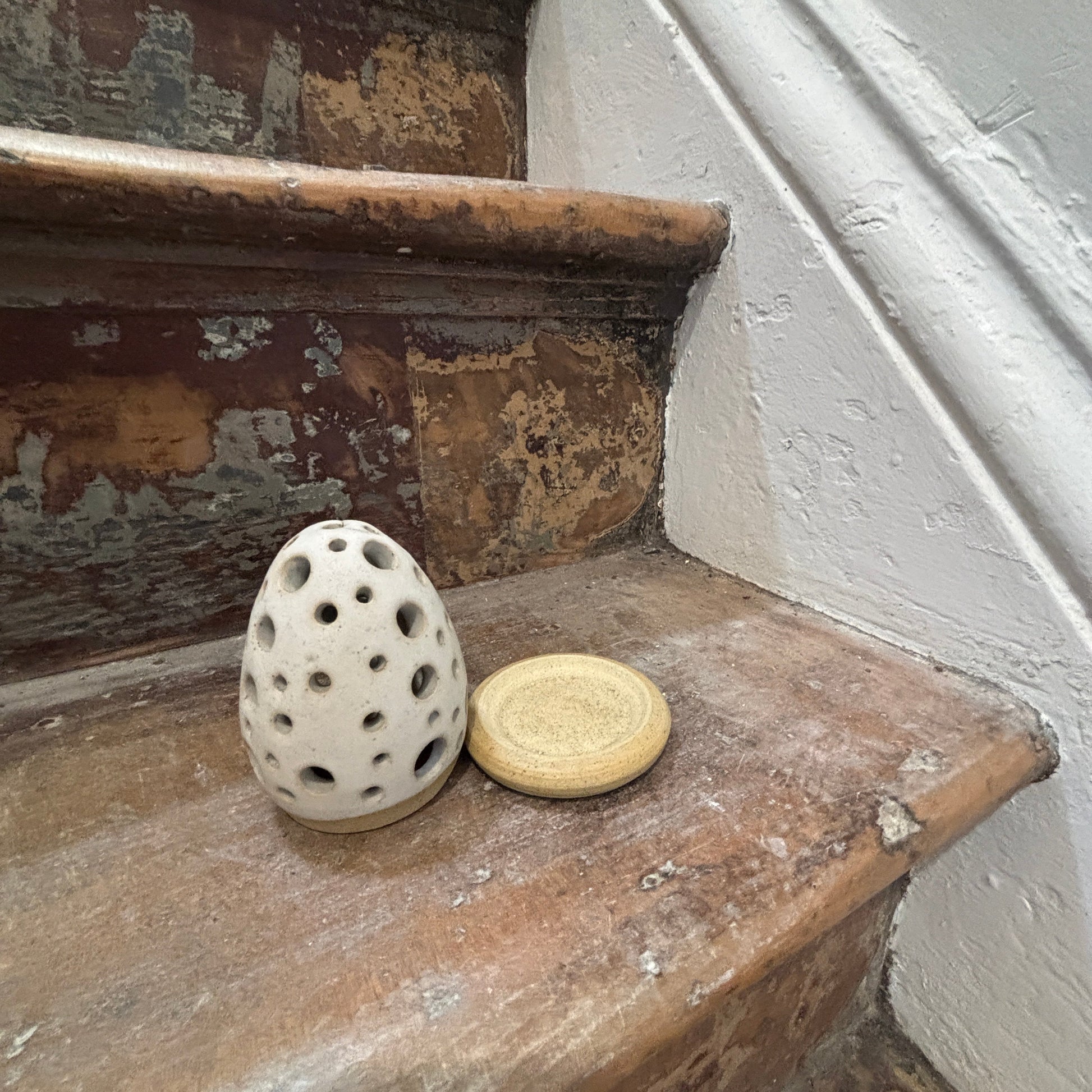 A small white ceramic egg-shaped incense holder sits on a weathered wooden staircase next to a round beige coaster or lid, with a white wall and stair railing in the background.