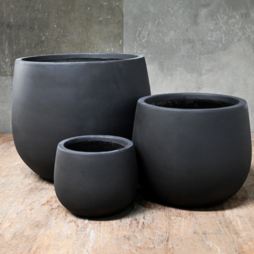 Three black planters of different sizes on a wooden surface with a gray concrete wall background.