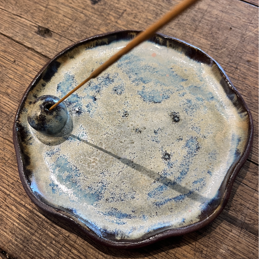 A handmade ceramic incense burner with a scalloped rim, featuring a textured beige and blue-gray glaze and a small protruding incense coil. A wooden incense stick is shown burning inside the burner, set on a rustic wooden tabletop.
