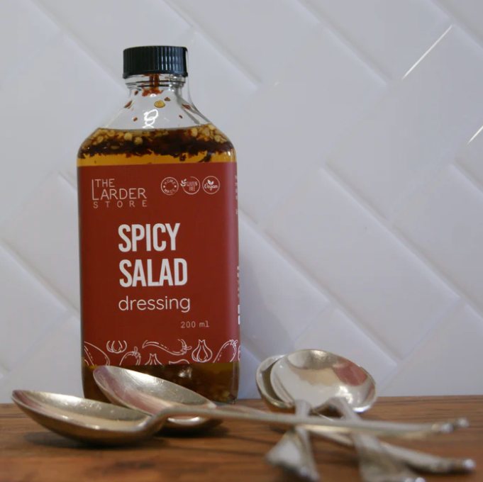 Bottle of 'Spicy Salad' dressing from 'The Larder' on a wooden surface with spoons.