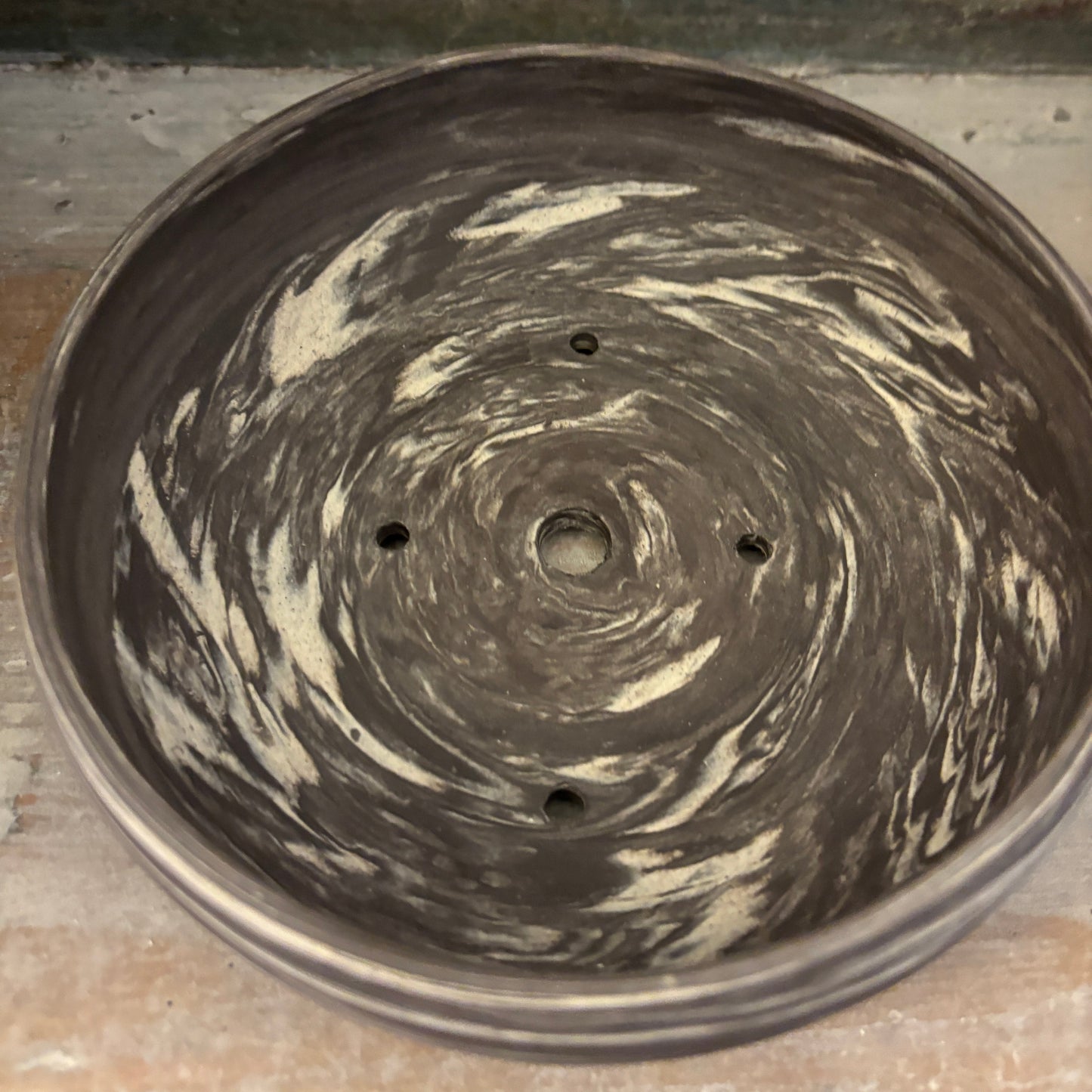 A round ceramic planter pot with a swirled, marbled pattern in dark charcoal gray and cream white, featuring drainage holes at the bottom, shown on a rustic wooden surface.