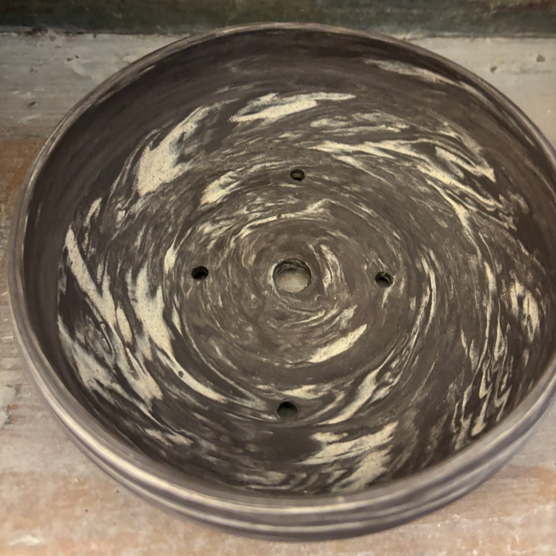 A round ceramic planter pot with a swirled, marbled pattern in dark charcoal gray and cream white, featuring drainage holes at the bottom, shown on a rustic wooden surface.