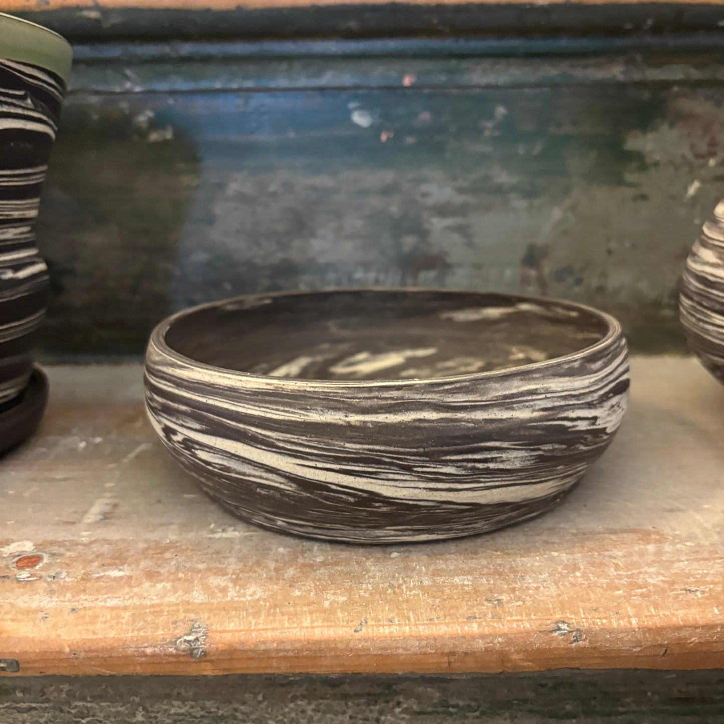 A handmade ceramic bonsai planter with a swirled black and white clay pattern, displayed on a rustic wooden shelf alongside other clay vessels.