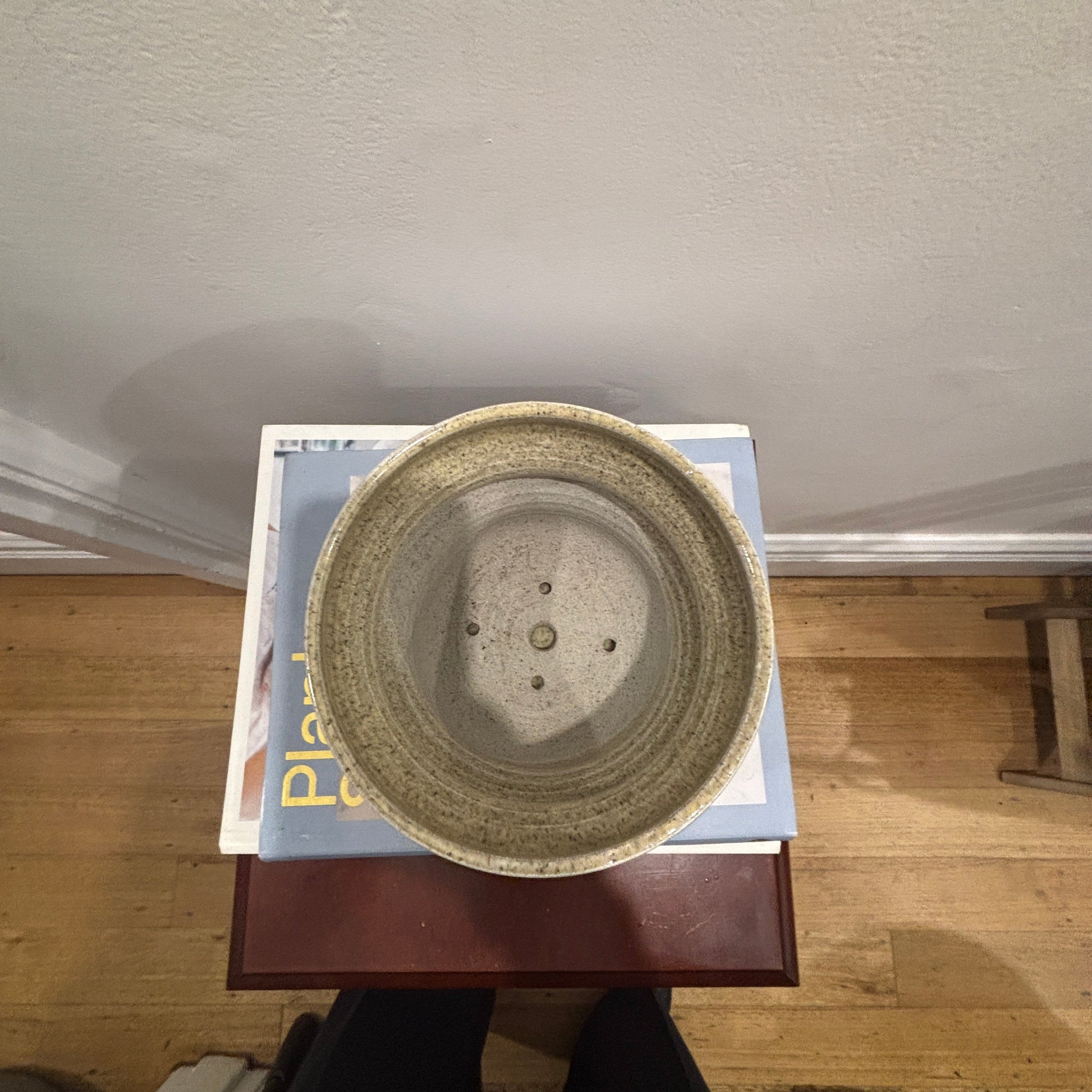 A handmade ceramic planter with a textured, oatmeal glaze, shown from a top-down perspective resting on a stack of books on a wooden floor.