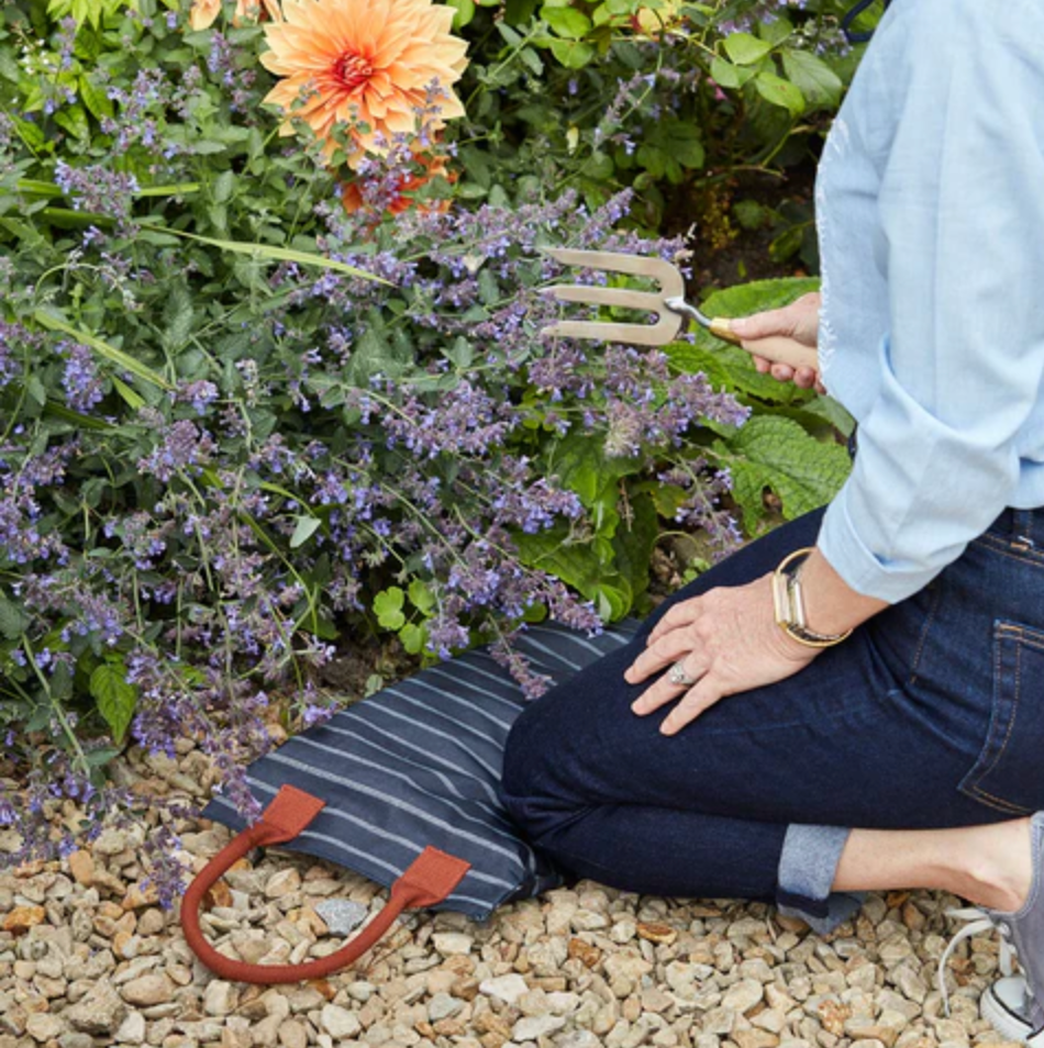 Person gardening with a striped garden cushion known as a kneeler and garden tool, surrounded by flowers and plants.