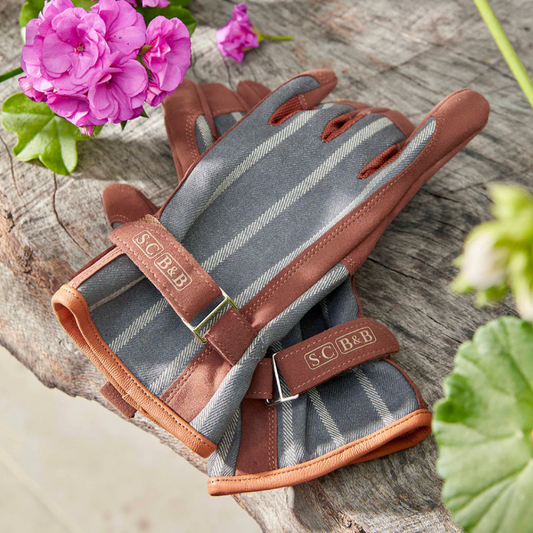 Pair of gardening gloves with brown leather straps on a wooden surface with flowers.