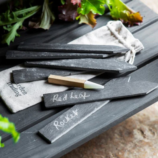 Set of slate garden labels with wooden handles on a wooden surface with plants in the background.