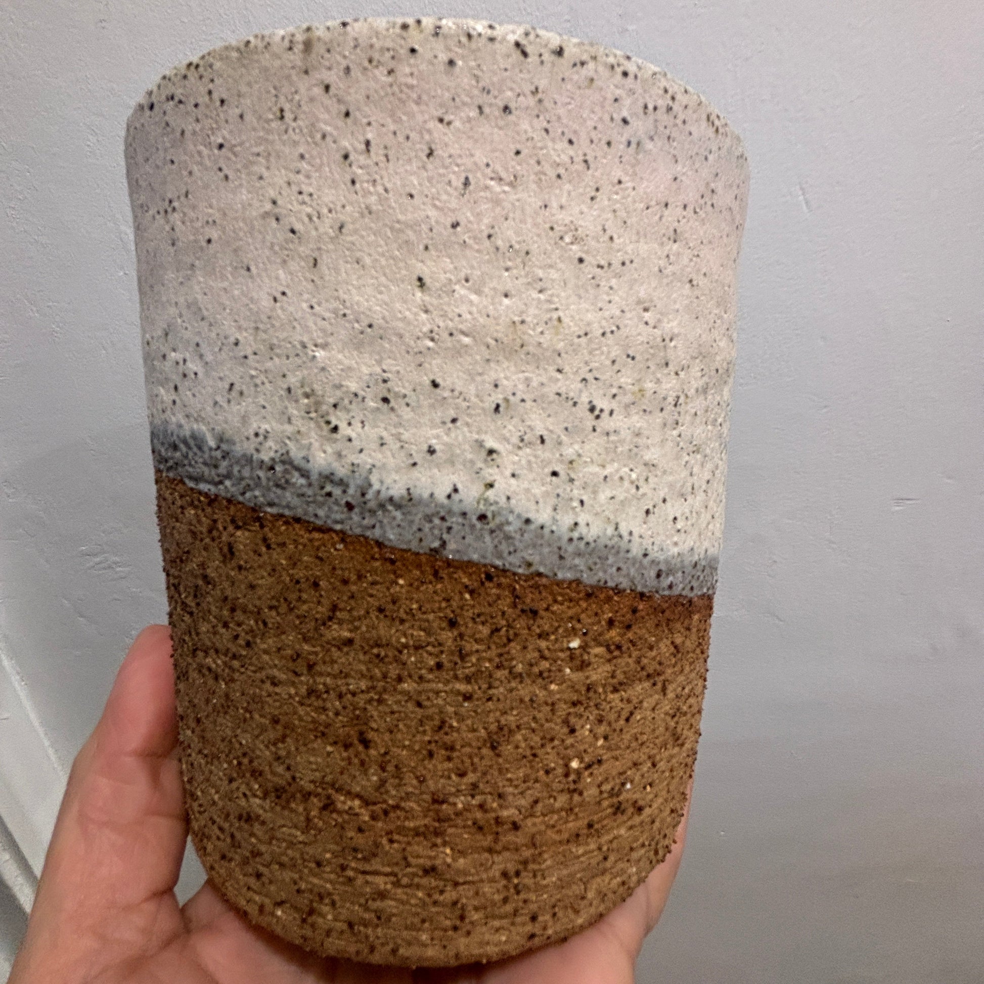 A handmade ceramic planter with a two-tone design, held in a hand against a plain white wall. The top half is speckled off-white with shades of pink and grey and the bottom half is a textured brown.