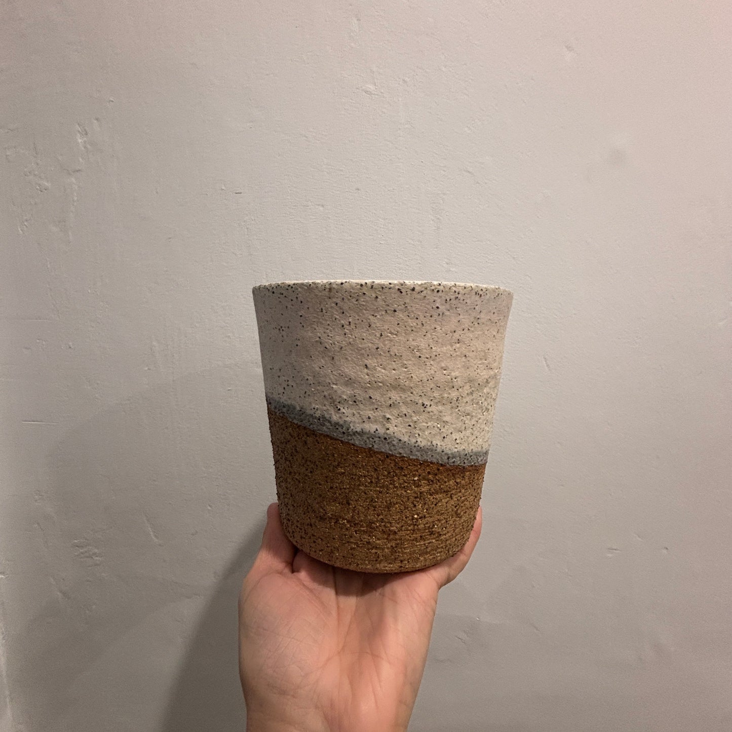 A handmade ceramic planter with a two-tone design, held in a hand against a plain white wall. The top half is speckled off-white with shades of pink and grey and the bottom half is a textured brown.