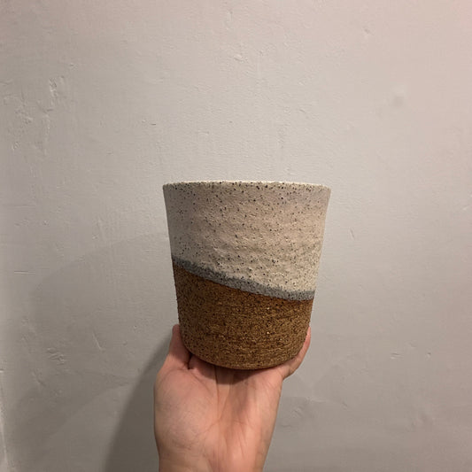 A handmade ceramic planter with a two-tone design, held in a hand against a plain white wall. The top half is speckled off-white with shades of pink and grey and the bottom half is a textured brown.