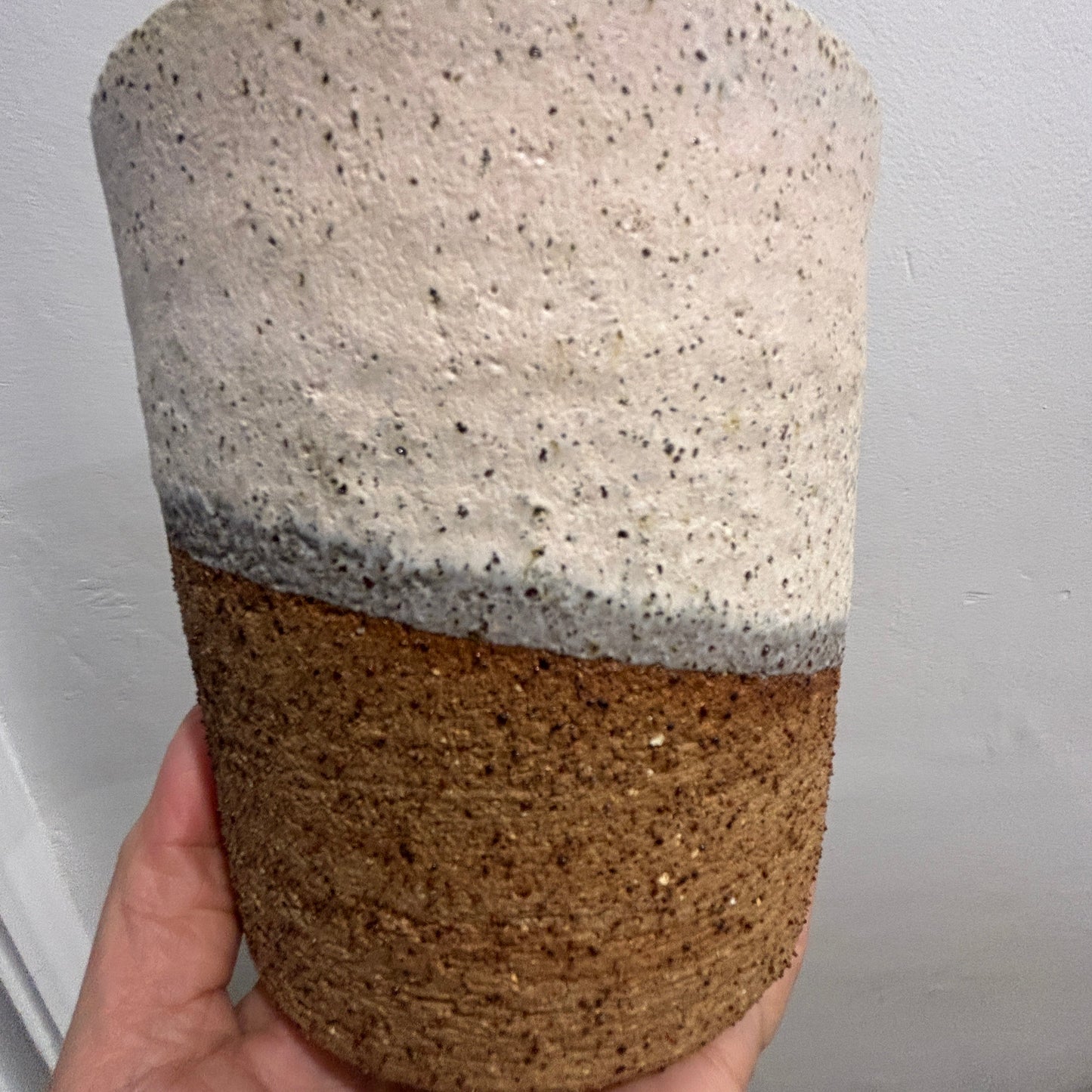A handmade ceramic planter with a two-tone design, held in a hand against a plain white wall. The top half is speckled off-white with shades of pink and grey and the bottom half is a textured brown.