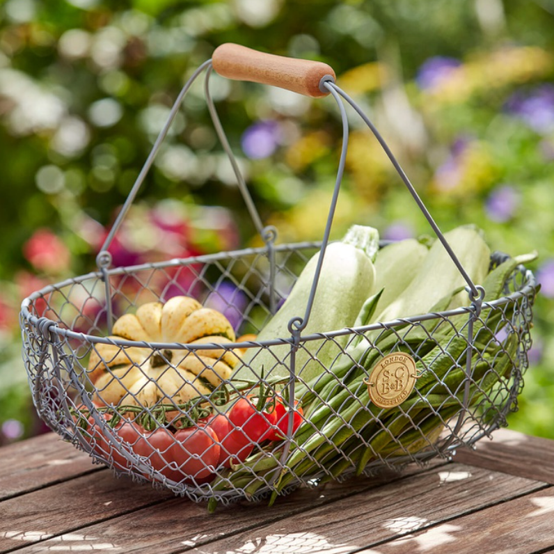 A gray wire garden basket with a natural wood handle, filled with fresh vegetables including zucchini, tomatoes, and a small gourd, set on a wooden deck with a blurred garden background.