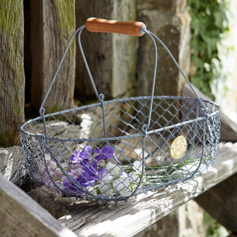 A rustic blue-gray wire basket with a curved wooden handle, resting on a weathered wooden step in front of a stone wall. Purple flowers are tucked inside the basket, and a small gold circular badge is attached to the side.