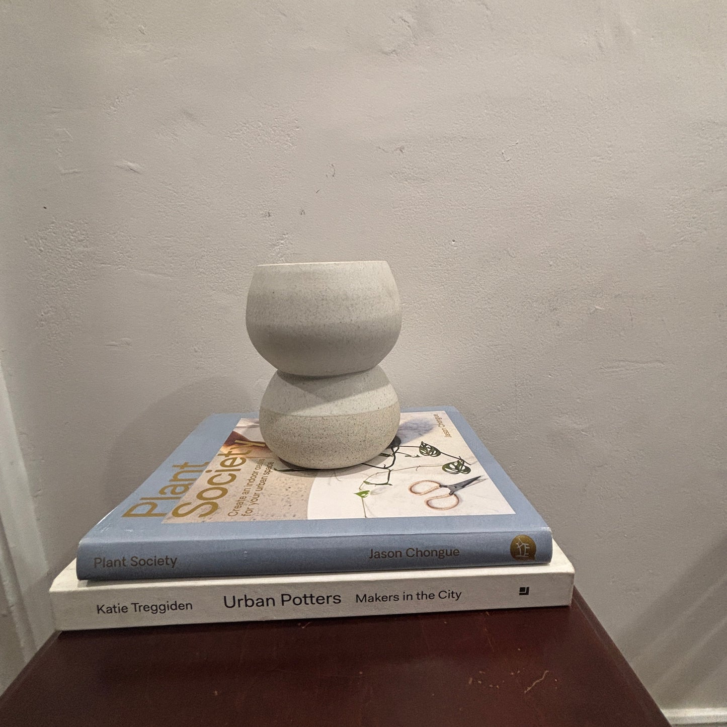 A small matte ceramic vase in a light grey color, styled on top of two books on a dark brown wooden side table against a textured off-white wall.