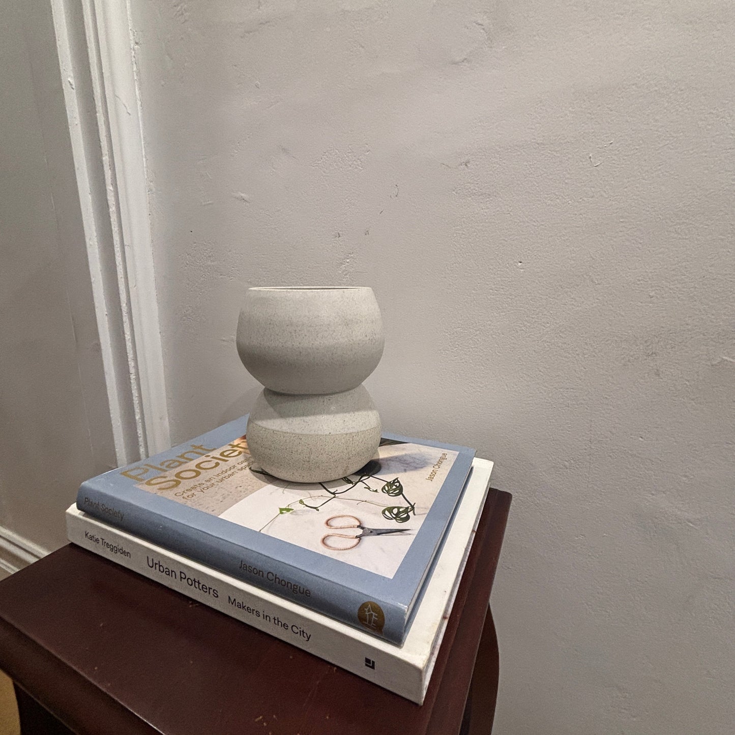 A small round stacked ceramic vase vessel sits on top of two books on a dark wooden side table against a textured light grey wall.